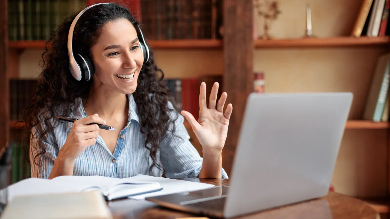 Student at desk with headphones and laptop, waiving to the computer screen.