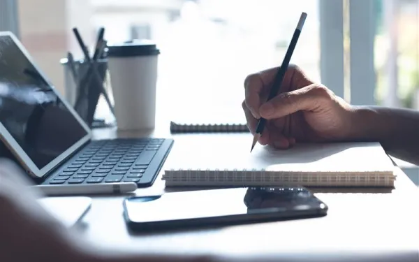 A person's hand writes in a notebook in front of a laptop.